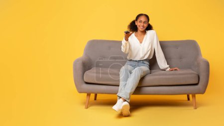 Photo for Black teenager girl in casual watching TV sitting on comfortable couch with remote television controller against yellow backdrop. Teen lady switches channels and watches film, empty space, panorama - Royalty Free Image