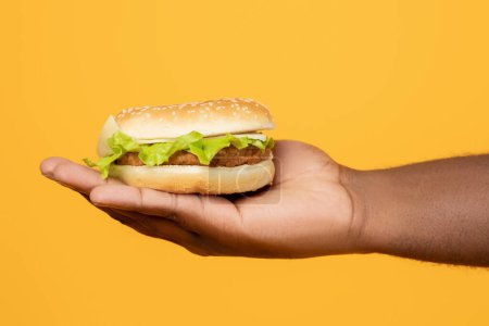 Photo for Junk Food. Cropped shot of black male hand holding tasty burger, offering unhealthy tasty snack, posing over yellow studio background. Closeup of guy showing cheeseburger, cheat meal - Royalty Free Image
