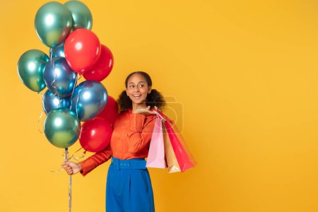 Photo for Holiday sales and shopping. Smiling black teen shopper girl holding bunch of bright balloons and paper shop bags, advertising discount over yellow studio background. Empty space for text - Royalty Free Image