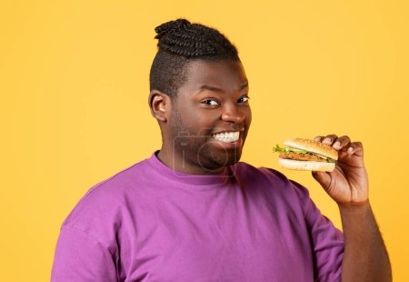 Téléchargez les photos : Homme noir souriant tenant un gros hamburger savoureux à la main, souriant à la caméra debout sur fond de studio jaune. Portrait d'un mangeur de malbouffe posant avec hamburger, publicité malbouffe - en image libre de droit