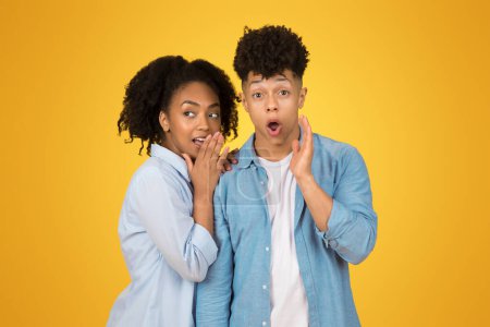 Photo for A young african american woman whispers into her hand with a mischievous expression while a young man next to her reacts with a hand to his cheek, both against a yellow background - Royalty Free Image