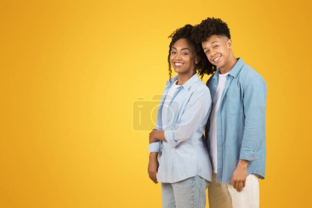 Photo for Two happy young woman and man with curly hair, wearing casual blue shirts, stand back to back with big smiles on vibrant yellow background, studio. Love, relationship, ad and offer - Royalty Free Image