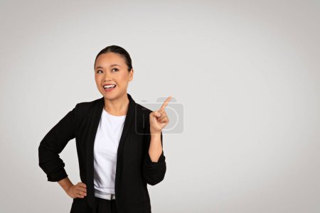 Téléchargez les photos : Femme d'affaires japonaise millénaire enthousiaste avec un sourire rayonnant pointant vers le haut, montrant optimisme et inspiration dans un cadre professionnel avec un arrière-plan neutre - en image libre de droit