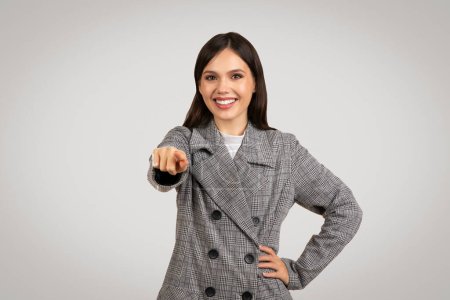 Photo for Cheerful young woman in fashionable plaid blazer, confidently pointing at the camera with a beaming smile, embodying positivity and directness - Royalty Free Image