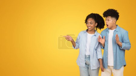 Foto de Una joven sorprendida y un hombre con atuendo casual señalan con entusiasmo algo fuera de cámara, con expresiones de sorpresa y deleite, contra un fondo amarillo brillante - Imagen libre de derechos