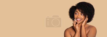 Téléchargez les photos : Femme afro-américaine rayonnante aux cheveux afro naturels souriant chaleureusement, touchant doucement son visage des deux mains, regardant de côté l'espace libre sur fond beige, panorama - en image libre de droit