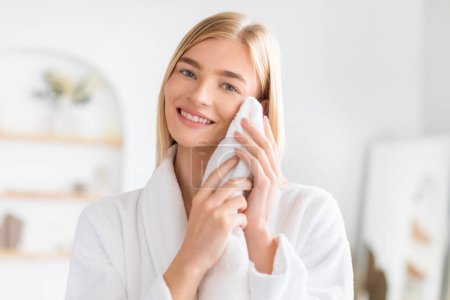 Téléchargez les photos : Jeune dame blonde souriante séchant son visage propre avec une serviette douce après avoir lavé à l'intérieur, debout dans un peignoir à la salle de bain moderne à l'intérieur, bénéficiant d'une hygiène faciale et de soins de la peau - en image libre de droit