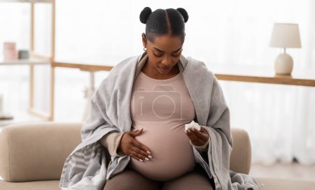 Photo for Seasonal flu. Pregnant young african american woman covered in blanket suffering from snotty nose, sitting on couch at home, holding napkin and touching her big tummy, empty space - Royalty Free Image