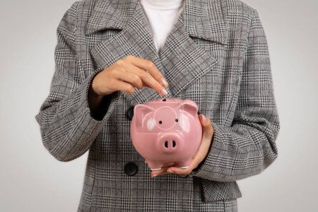 Photo for Closeup of woman in checkered blazer depositing coin into pink piggy bank, symbolizing savings and financial planning, against a grey background - Royalty Free Image