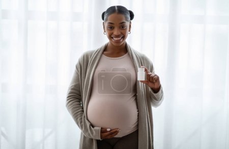 Téléchargez les photos : Femme enceinte afro-américaine positive montrant un pot en plastique blanc avec des vitamines, des suppléments pour les femmes enceintes, debout à côté de la fenêtre à la maison, étreignant son gros ventre - en image libre de droit