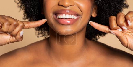 Foto de Primer plano de mujer negra radiante con el pelo rizado apuntando a sus dientes blancos brillantes con expresión alegre, colocada sobre fondo beige, recortada - Imagen libre de derechos