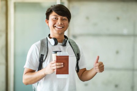 Téléchargez les photos : Heureux jeune homme asiatique excité tenant passeport et carte d'embarquement ou billets, montrant pouce levé et souriant à la caméra, portant un sac à dos, voyageant à l'étranger, espace de copie - en image libre de droit