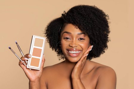 Photo for Radiant woman with afro hair joyfully holding a makeup palette and brushes, displaying tools for beauty enhancement against beige background - Royalty Free Image