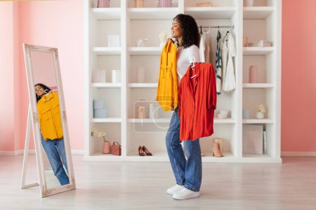 Photo for Cheerful woman with curly hair trying on a yellow and holding a red shirt, making choice in front of a mirror in her bright room, full length - Royalty Free Image