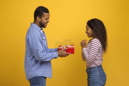 Photo for Happy young black man giving red gift box to his delighted girlfriend, romantic african american guy greeting wife with birthday or valentines day, standing against bright yellow background - Royalty Free Image
