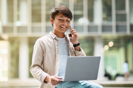 Téléchargez les photos : Millennial homme d'affaires asiatique entrepreneur assis sur le banc au centre d'affaires, en utilisant un ordinateur portable et parler au téléphone avec le client, multitâche dans le concept d'entreprise, espace de copie - en image libre de droit
