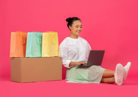 Téléchargez les photos : Jeune femme asiatique assise avec ordinateur portable, entourée de sacs à provisions colorés, heureuse coréenne profitant d'une expérience de magasinage en ligne transparente, se détendre sur fond rose vif studio - en image libre de droit
