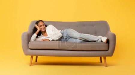 Téléchargez les photos : Adolescente afro-américaine insouciante se détendant allongée sur un canapé et souriant à la caméra, profitant du confort et de son repos sur fond de studio jaune. Concept de loisirs de détente. Panorama - en image libre de droit
