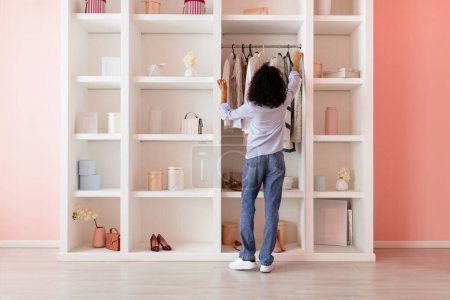 Photo for Curly-haired woman in casual attire thoughtfully organizing her wardrobe in a modern, neatly arranged closet in a room with pink walls, back view - Royalty Free Image