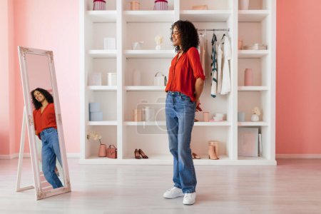 Téléchargez les photos : Femme souriante portant un chemisier rouge et un jean bleu, admirant son reflet dans un miroir pleine longueur dans une pièce aux murs roses et aux étagères blanches - en image libre de droit