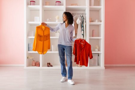 Photo for Happy curly-haired latin woman holding yellow and red shirts, comparing them for her outfit in a chic closet within a room with pastel pink walls - Royalty Free Image
