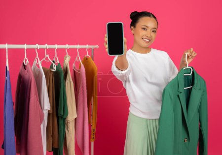 Photo for Fashion App. Smiling asian fashionista woman showing blank smartphone screen and holding stylish green blazer, happy lady recommending app or website, standing near clothing rack on pink background - Royalty Free Image