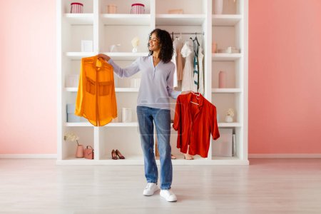 Photo for Curly-haired woman happily comparing vibrant orange shirt with red one as she organizing her fashionable wardrobe in cozy pink room, full length - Royalty Free Image
