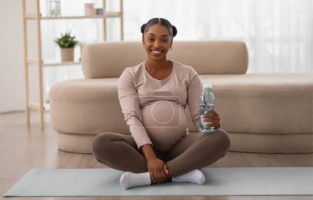 Téléchargez les photos : Heureuse belle jeune femme enceinte afro-américaine assise sur un tapis de yoga, tenant une bouteille, de l'eau potable après l'entraînement à la maison. Fitness, mode de vie sain pour les femmes enceintes - en image libre de droit