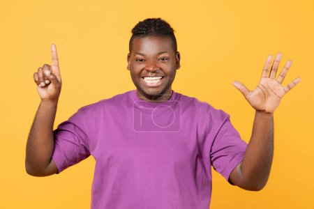 Téléchargez les photos : Toothy Afro-Américain levant les deux mains montrant le numéro six, paumes ouvertes avec cinq et un doigt sur fond de studio jaune, comptant jusqu'à six, souriant à la caméra - en image libre de droit