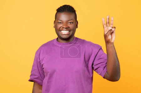 Foto de Alegre chico afroamericano en camiseta púrpura levantando la mano mostrando tres dedos, contando con gesto, sonriendo a la cámara mientras posando sobre el fondo del estudio amarillo, retrato disparado - Imagen libre de derechos