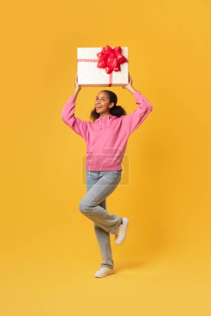 Foto de Tiro vertical de adolescente negro feliz levantando brazos sosteniendo una gran caja de regalo con lazo de cinta sobre el fondo amarillo del estudio. Oferta regalo de cumpleaños, concepto de celebración de vacaciones - Imagen libre de derechos