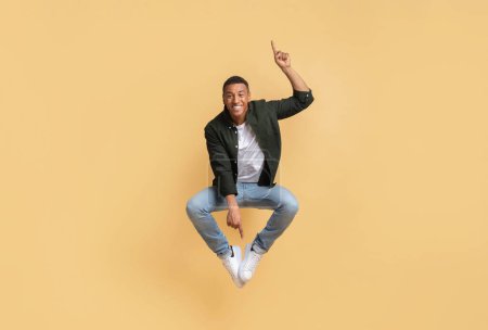 Photo for Emotional handsome young african american guy in stylish casual outfit hipster jumping in the air, pointing up and down, isolated on beige colored background, copy space - Royalty Free Image