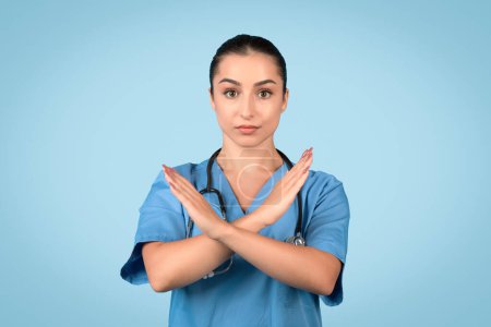 Téléchargez les photos : Infirmière européenne sérieuse en uniforme bleu croisant les bras pour former un signe X, symbolisant refus ou interdiction, posant sur un fond bleu propre - en image libre de droit