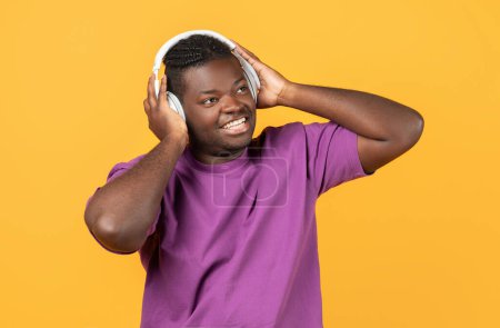 Foto de Música favorita. Hombre negro alegre en camiseta púrpura casual con auriculares inalámbricos, escucha una gran canción en línea, mirando a un lado con sonrisa sobre el fondo del estudio amarillo - Imagen libre de derechos