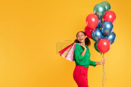 Photo for Birthday sales. Cheerful black teenage shopper girl carries colorful balloons and paper shopping bags, embodying the spirit of holiday celebration and shop spree against yellow backdrop. Free space - Royalty Free Image