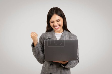 Photo for Smiling businesswoman in checkered suit holding laptop and making triumphant fist pump, celebrating victory or success, on light grey background - Royalty Free Image