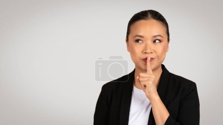 Photo for Asian businesswoman with a finger on her lips making a silence gesture, conveying a sense of secrecy or the need for quiet in a professional setting, isolated on gray background, studio - Royalty Free Image