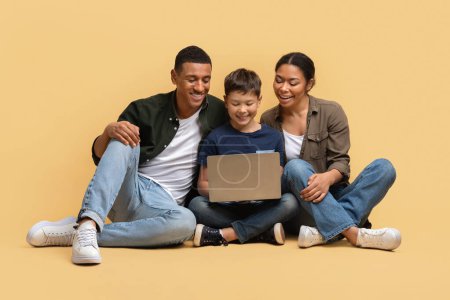Photo for Beautiful family of three father, mother and school-aged son sitting on floor over beige studio background, watching movie or cartoon together, websurfing, using laptop computer - Royalty Free Image
