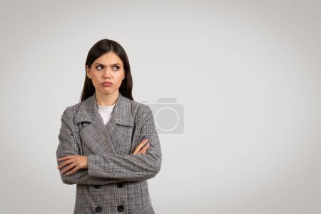 Téléchargez les photos : Jeune femme d'expression sceptique portant une veste en tweed à dents de poule, bras croisés, regardant de côté l'espace libre, montrant un sentiment de doute ou de pensée critique - en image libre de droit