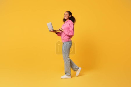 Téléchargez les photos : Étudiante noire souriante utilisant un ordinateur portable pour l'éducation marchant sur fond jaune en studio. Plan complet de jeune adolescent naviguant sur le web en déplacement, espace libre pour l'offre éducative en ligne - en image libre de droit