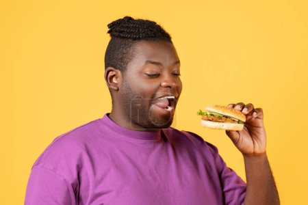 Téléchargez les photos : Délicieux Fast Food. Gros plan portrait de afro-américain affamé mangeant burger, tenant et mordant savoureux malbouffe sur fond jaune orangé studio. Repas Cheat malsain - en image libre de droit