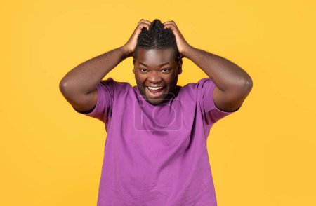 Téléchargez les photos : Homme afro-américain émotionnel en t-shirt violet criant fort touchant la tête et regardant la caméra, posant sur fond de studio jaune, prise de vue portrait. Concept d'Omg - en image libre de droit