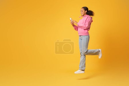 Foto de Feliz estudiante afroamericana salta en el aire navegando por su teléfono inteligente, encapsulando la juventud y la tecnología en el fondo amarillo, navegar por Internet en gadget. Longitud completa, espacio libre - Imagen libre de derechos