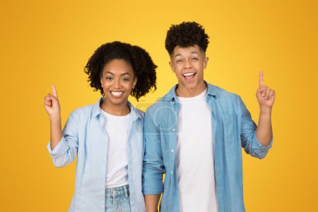 Photo for Happy and confident african american young woman and man with cheerful smiles pointing upwards, standing together against an upbeat yellow background, exuding optimism and enthusiasm - Royalty Free Image