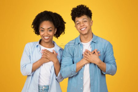 Téléchargez les photos : Joyeux jeune femme afro-américaine et homme aux expressions joyeuses, les mains sur le cœur, debout ensemble devant un fond jaune chaud, exsudant positivité et amitié - en image libre de droit