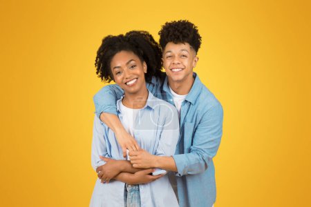 Photo for A radiant glad young woman and man in casual blue shirts embrace warmly, sharing a genuine smile, with the mans arm around her on a bright yellow background. Love, relationship, ad and offer - Royalty Free Image