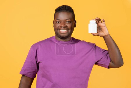 Téléchargez les photos : Homme noir joyeux en t-shirt violet montrant pot de pilules à la caméra et souriant, debout sur fond de studio jaune. Recommandation médicale, suppléments et concept de traitement thérapeutique - en image libre de droit