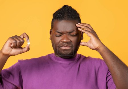 Photo for African american man having painful headache holding painkiller pill and touching head, posing on yellow background in studio, suffering from pain, coping with severe migraine symptoms - Royalty Free Image