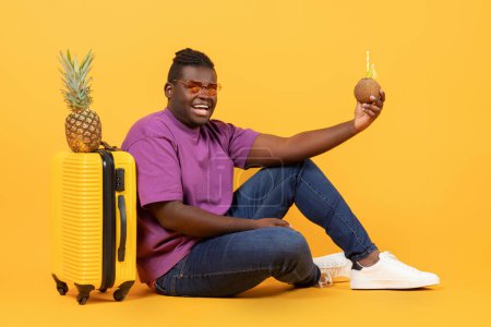 Téléchargez les photos : Offre vacances d'été. Un noir excité assis près d'une valise contenant de l'ananas et de la noix de coco avec des pailles, prêt à voyager dans un pays exotique, posant sur un fond de studio jaune - en image libre de droit
