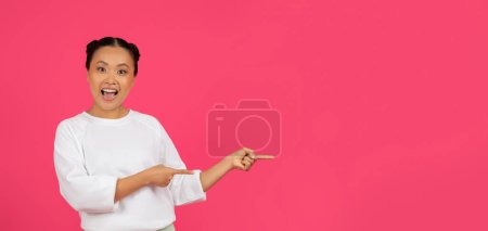 Foto de Bonita oferta. Emocionado joven asiática mujer señalando a un lado con ambas manos, feliz alegre coreana mujer demostrando espacio de copia para la publicidad, de pie sobre fondo rosa brillante, panorama - Imagen libre de derechos